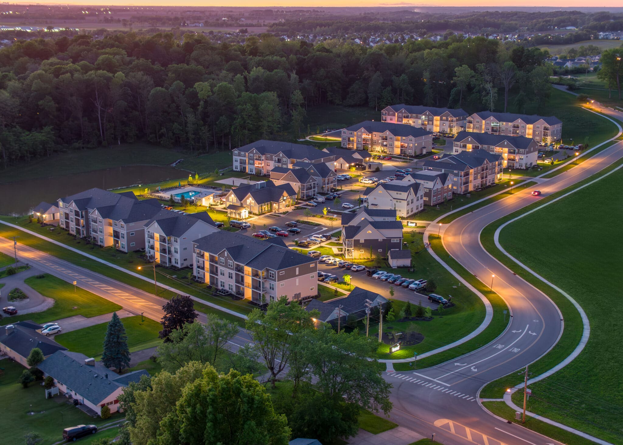 An aerial view of a modern housing complex at dusk, showing multiple apartment buildings surrounded by well-lit streets and parking areas. The surrounding area includes lush green trees and a curved road leading into the complex, with the sun setting in the distant horizon.